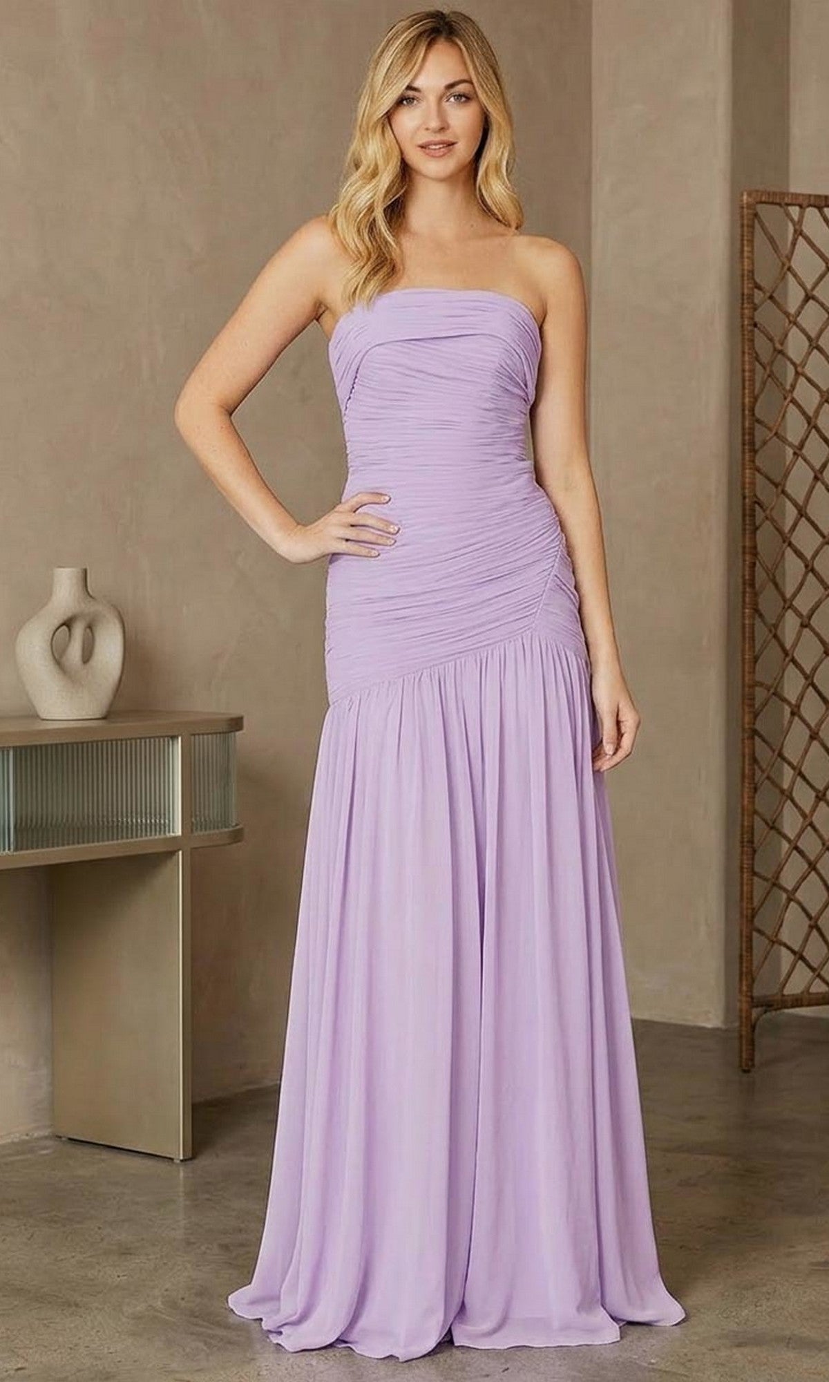 A woman with light wavy hair smiles slightly indoors while wearing the Amelia Couture 6132 Long Prom Dress in lilac, featuring a strapless neckline, floor-length silhouette, and figure-flattering ruching.